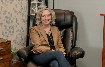 A woman with curly blonde hair sitting in a black leather chair in a room with patterned wallpaper and a wooden side table.