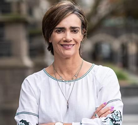 A woman with short brown hair smiling, wearing a white long-sleeve shirt with embroidery, standing outdoors with a blurred background.