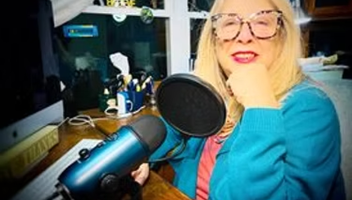 A woman with blonde hair and glasses sitting at a desk in front of a computer, holding a blue microphone, smiling at the camera.