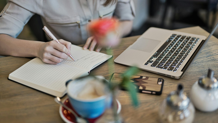 Person writing in notebook at a wooden table with a laptop, smartphone, and tea cup, with blurred flowers in the foreground.