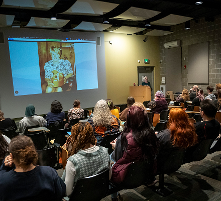 Audience attending a presentation in a conference room, watching a slideshow of a young girl holding a toy on a projector screen.