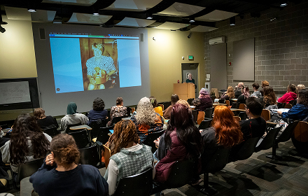Audience attending a presentation in a conference room with a speaker on a slideshow screen.
