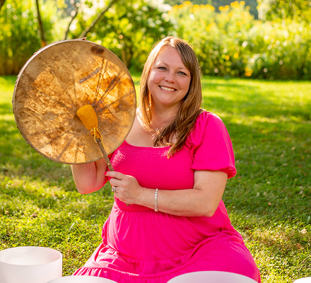 A woman in a pink dress smiling and holding a large drumstick outdoors in a park with green trees and grass.