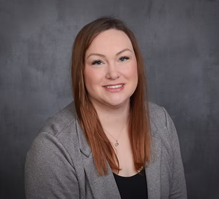 Portrait of a woman with shoulder-length red hair, wearing a gray blazer and black top, smiling against a gray background.