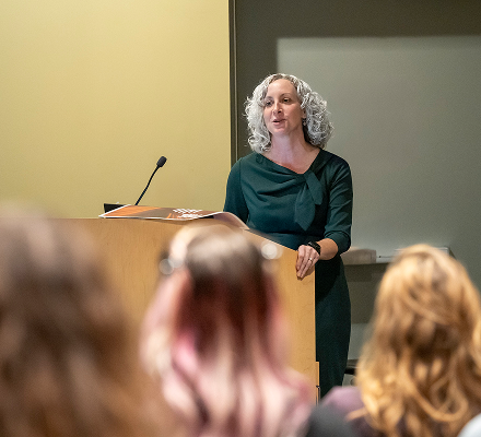 A woman with curly gray hair standing at a podium giving a presentation in a lecture hall.