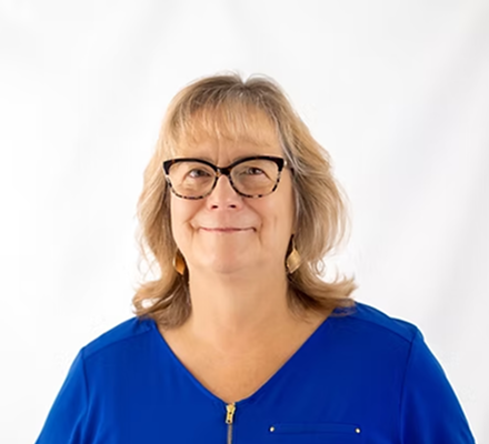A woman with blonde hair, glasses, and a blue top standing against a white background.
