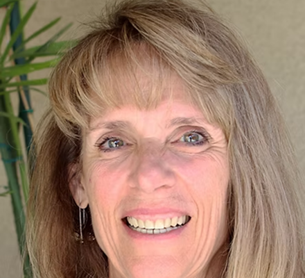 A woman with blonde hair smiling, standing in front of a green plant.