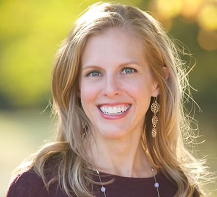 A smiling woman with blonde hair and blue eyes outside, wearing earrings and a dark top, with a blurred green and yellow background.