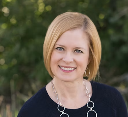 A woman with short blonde hair smiling outdoors, wearing a navy sweater and a silver necklace with circular links.