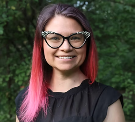 Young woman with glasses and pink-tinted hair smiling outdoors with green trees in the background.