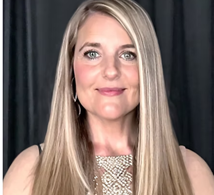 A woman with long blonde hair, wearing a black top and a pearl necklace, standing in front of a black curtain.