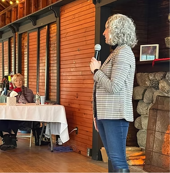 A woman with curly gray hair speaking into a microphone at an indoor event, with an audience member sitting at a table watching her.