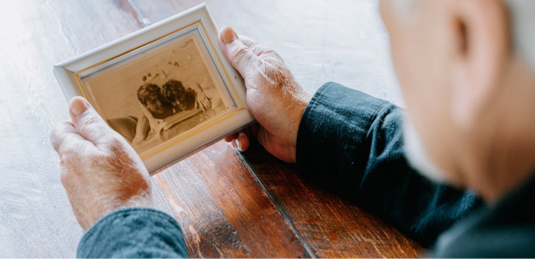 An elderly person holding a framed photograph of two children kissing, sitting at a wooden table.