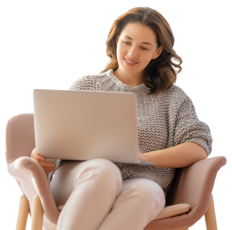 A young woman with wavy brown hair sitting in a pink armchair, working on a silver laptop, smiling and wearing a beige knitted sweater and light-colored pants.