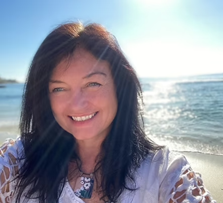 Woman smiling at the beach with sunlight shining on her and the ocean in the background.