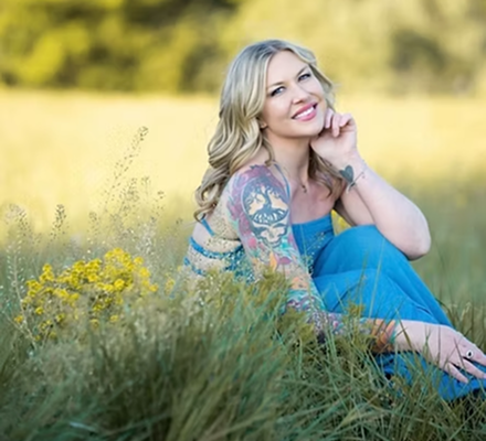 A woman with blonde hair and tattoos sitting on grass in a field, smiling at the camera.
