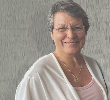 A smiling woman with short gray hair, wearing glasses, a light pink top, and a white cardigan, standing against a gray textured wall.