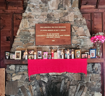 Memorial display on a stone fireplace with a plaque, framed photos, candles, and pink flowers.