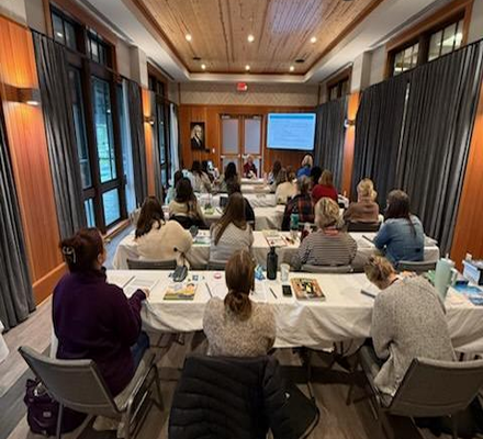A group of people in a meeting or conference room, seated at tables, watching a presentation on a screen at the front of the room.