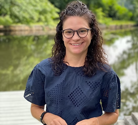 A woman with curly hair and glasses smiling outdoors near a body of water with trees in the background.