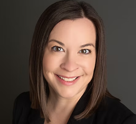 A woman with shoulder-length brown hair, smiling, wearing a black top, against a gray background.