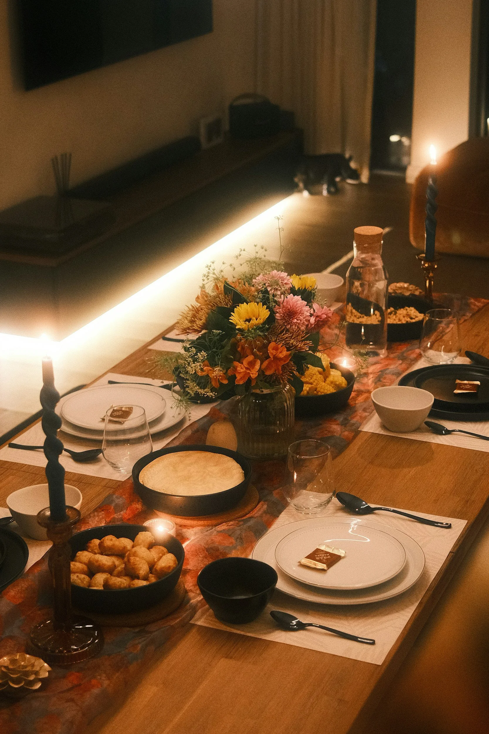 A warmly lit dining table set for a meal with a flower centerpiece, plates, glasses, and food including bread, snacks, and a jar of water, in a cozy room with candles and decor.