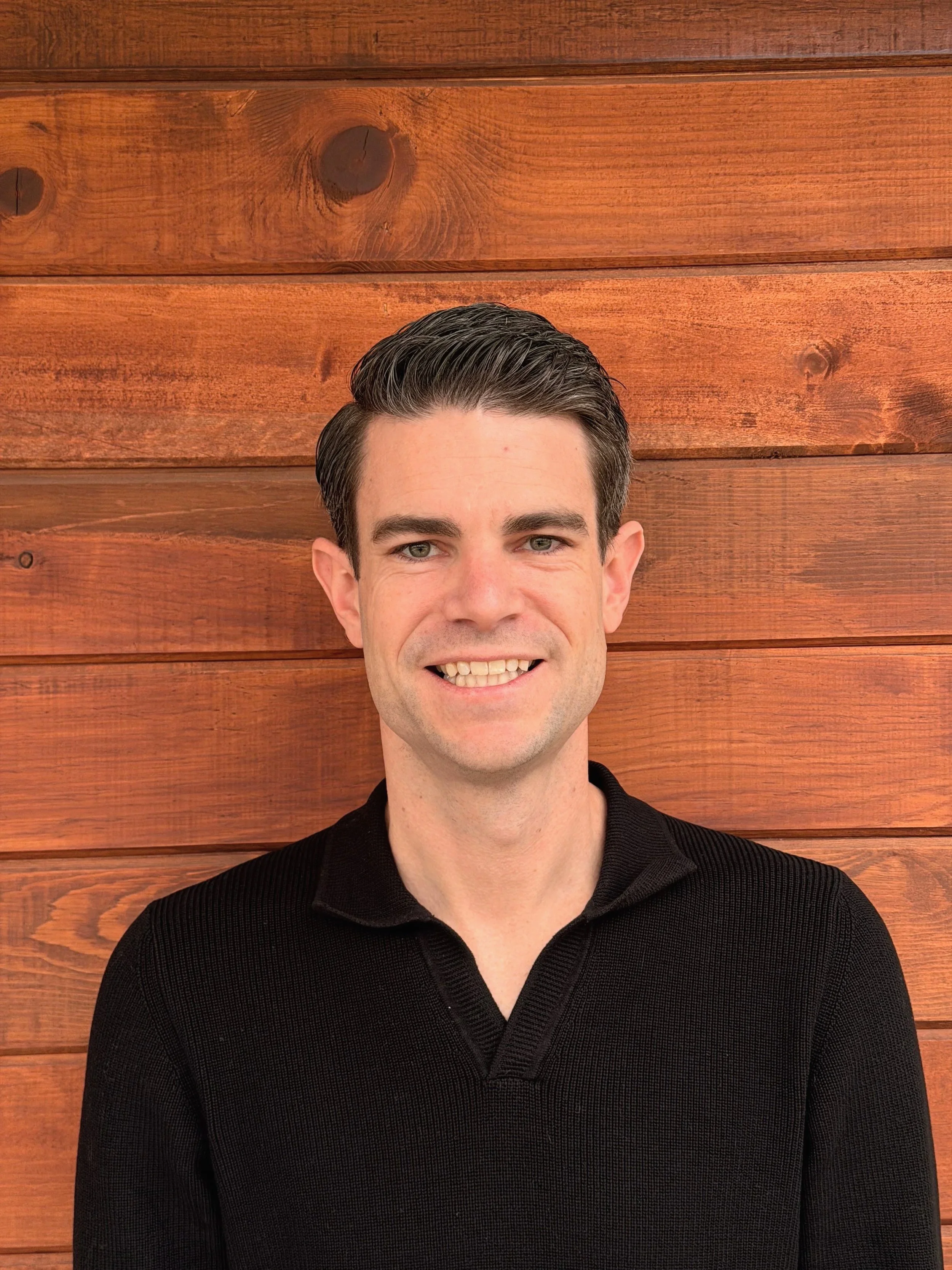 Young man with dark hair and a black collared shirt smiling in front of a wooden wall.