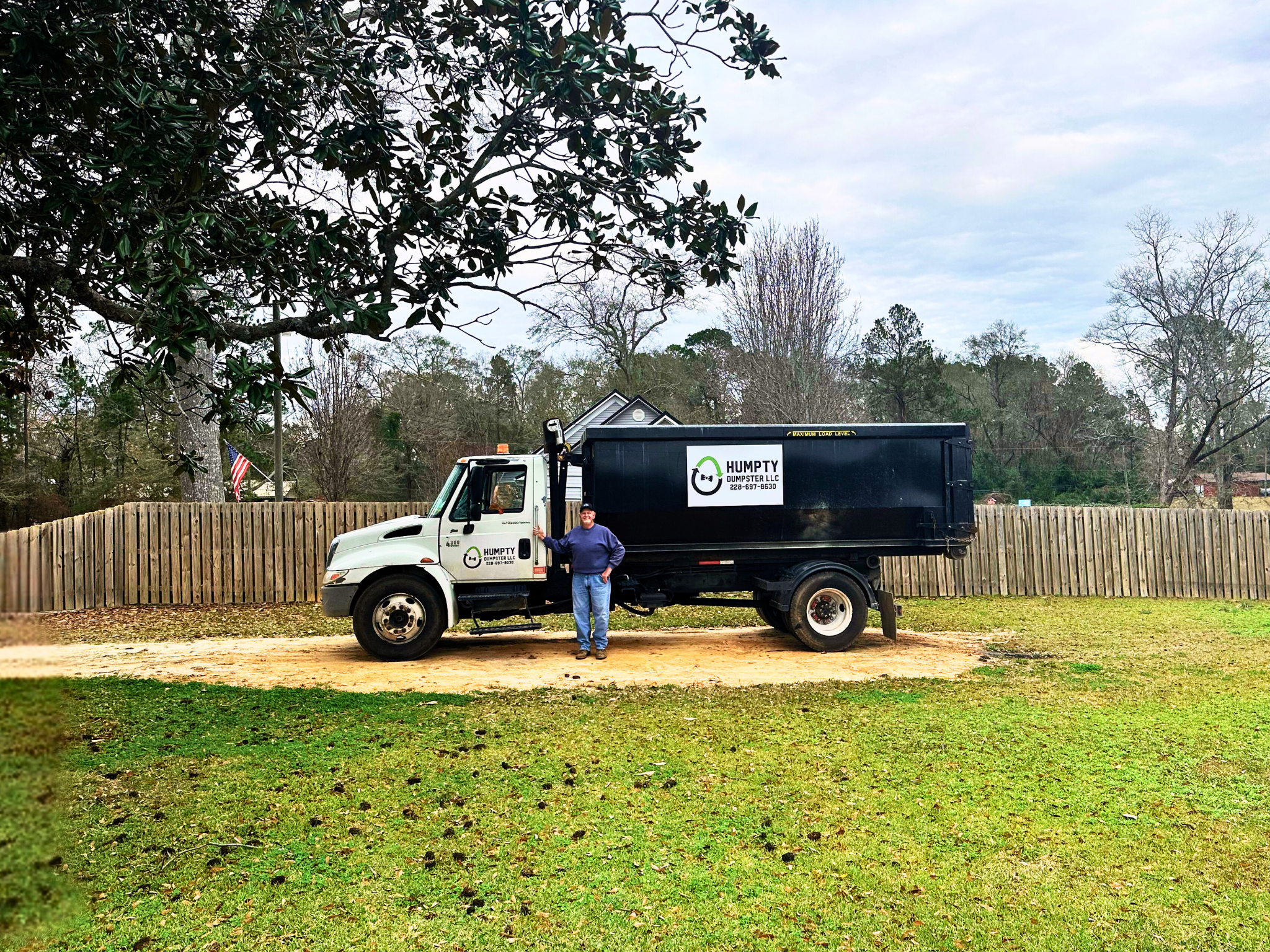 A man standing next to a black dumpster truck with a white cab, parked on a grassy area in front of a wooden fence. The truck has a sign that reads 'HUMPTY DUMPSTER LLC' with a phone number. There are trees and a house in the background under a cloudy sky.