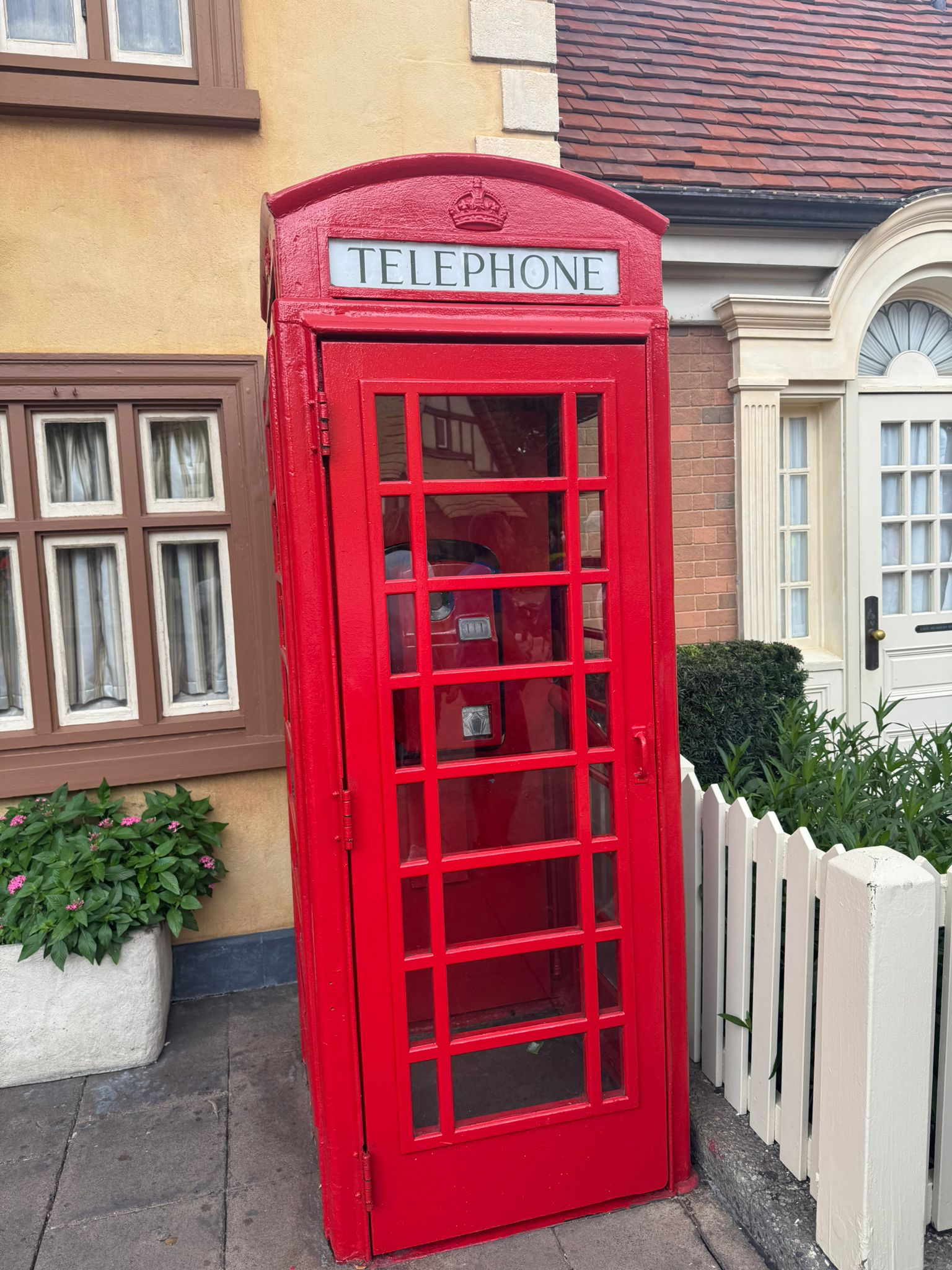 Red British-style telephone booth on a sidewalk in front of a house with beige and brick walls, white window frames, and a white picket fence. [Copyright - Photography by Tom]