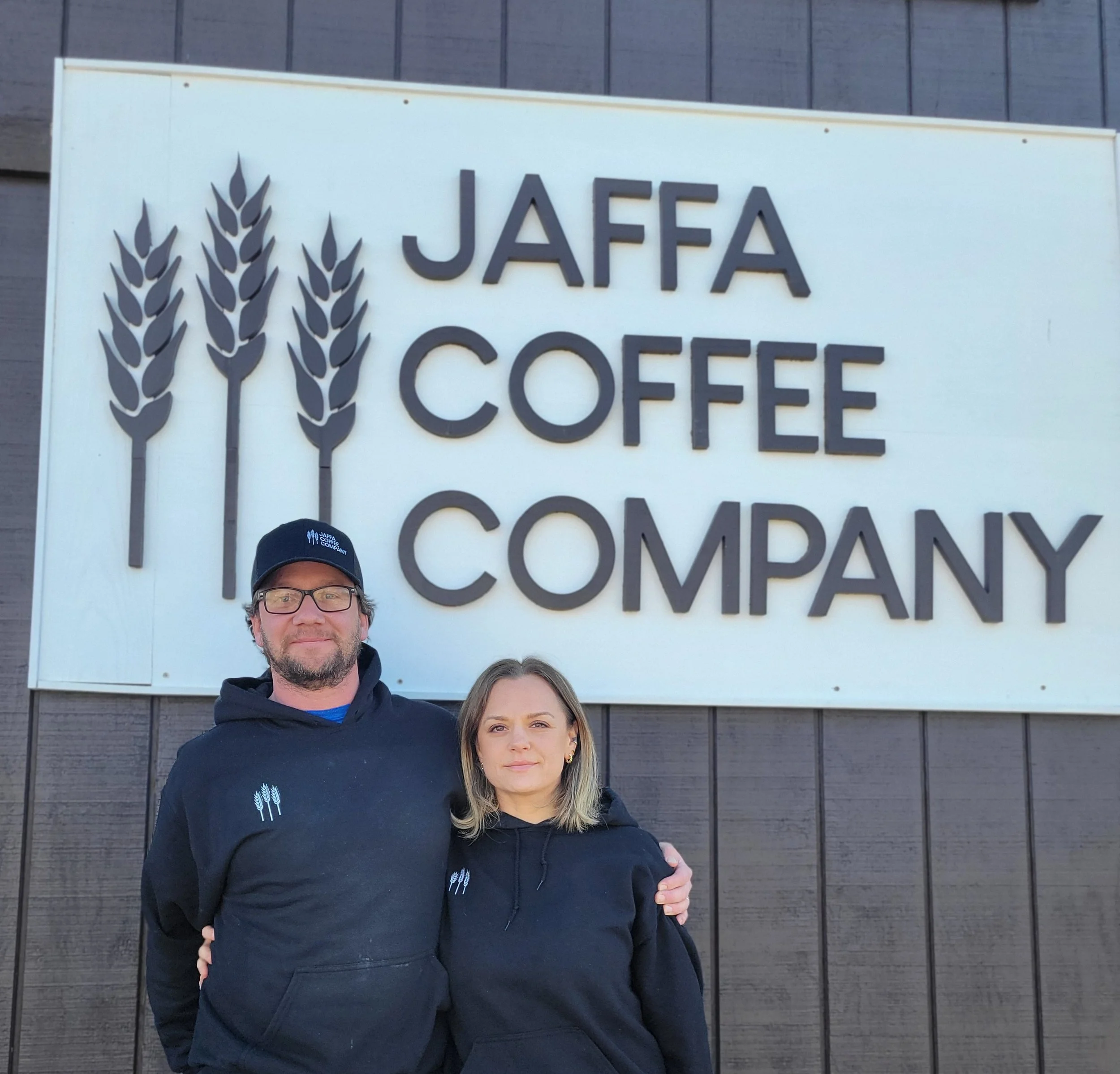 A man and a woman standing in front of a sign that reads 'JAFFA COFFEE COMPANY' with three wheat stalks graphic. They are both wearing black hoodies with a small wheat stalk logo and the man is wearing a black cap.