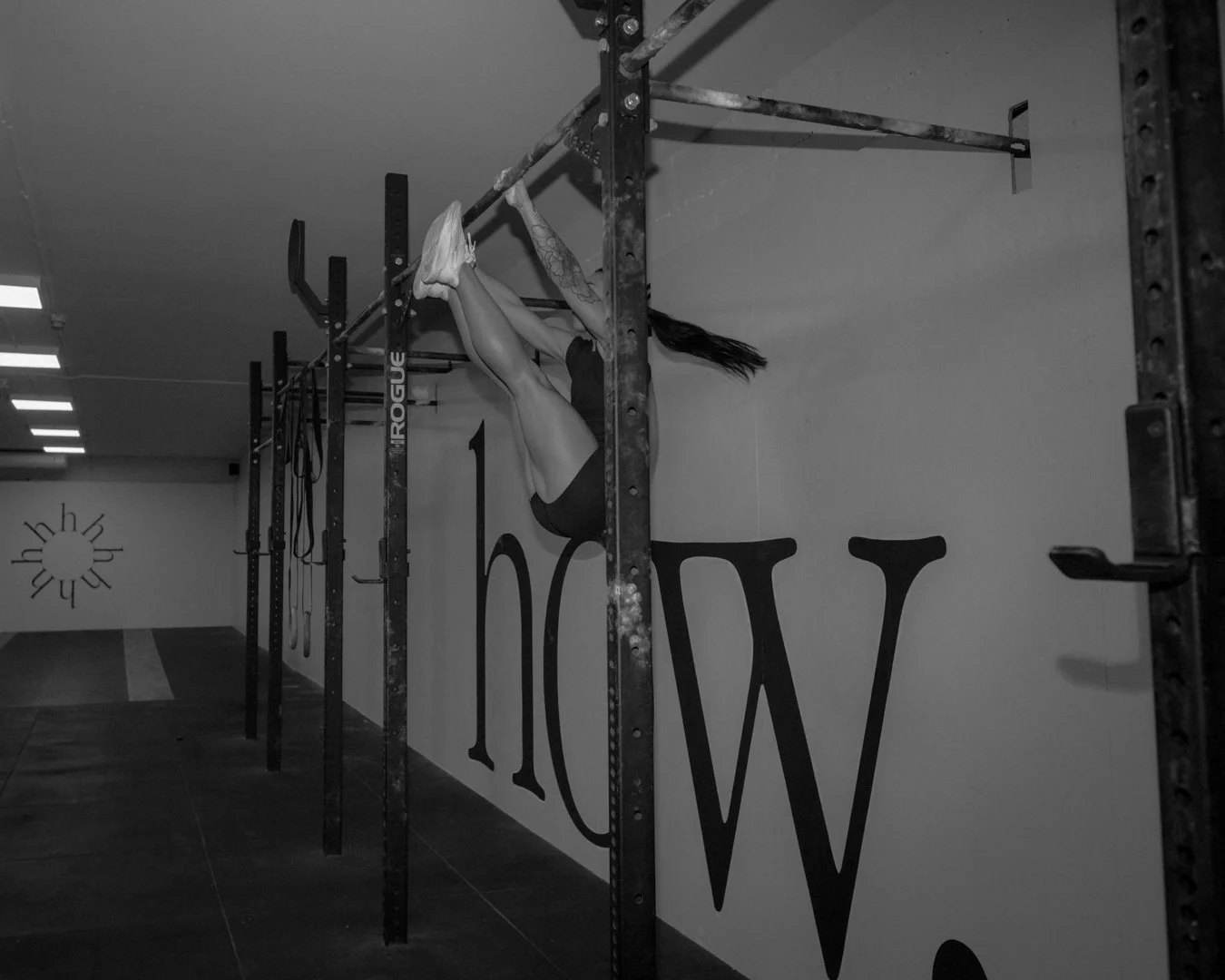 Una mujer hace ejercicio en un gimnasio, colgada de una barra, en la pared hay letras grandes formando la palabra 'how'.