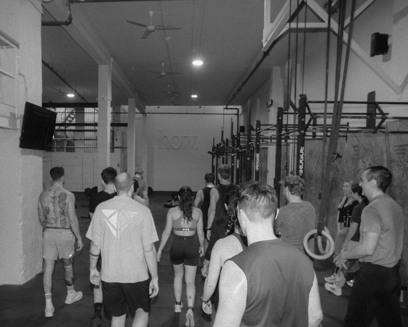 Grupo de personas en un gimnasio, algunas con ropa deportiva y otras sin camiseta, participando en una clase o entrenamiento con equipo de CrossFit, en un espacio con paredes blancas y equipo de entrenamiento en fondos.