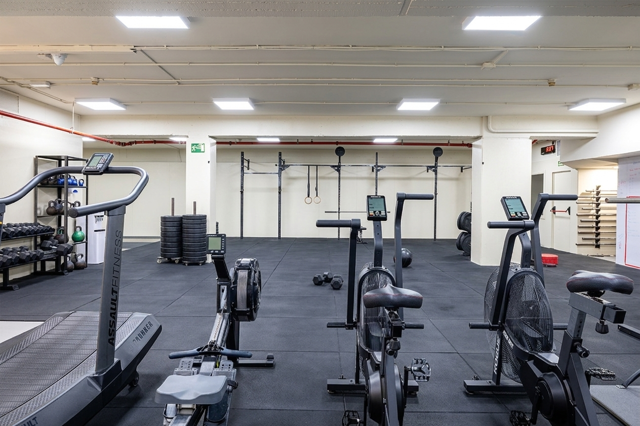 Gimnasio vacío con equipos de cardio y pesas, paredes blancas y piso de goma negra.