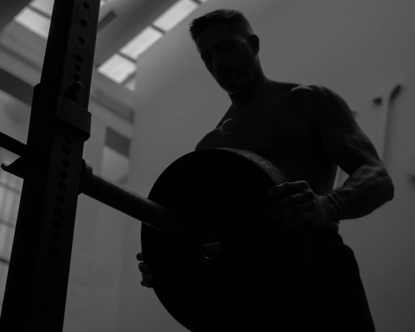 Persona en un gimnasio con sombra, sosteniendo una pesa en la mano y una barra de pesas en un fondo con ventanas.