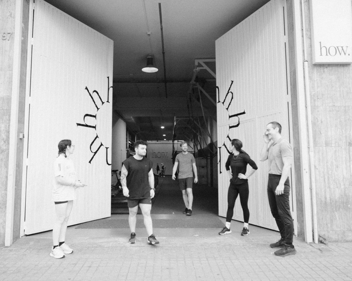 Grupo de cinco personas en la entrada de un gimnasio, algunas mirándose, en un entorno moderno y luminoso.