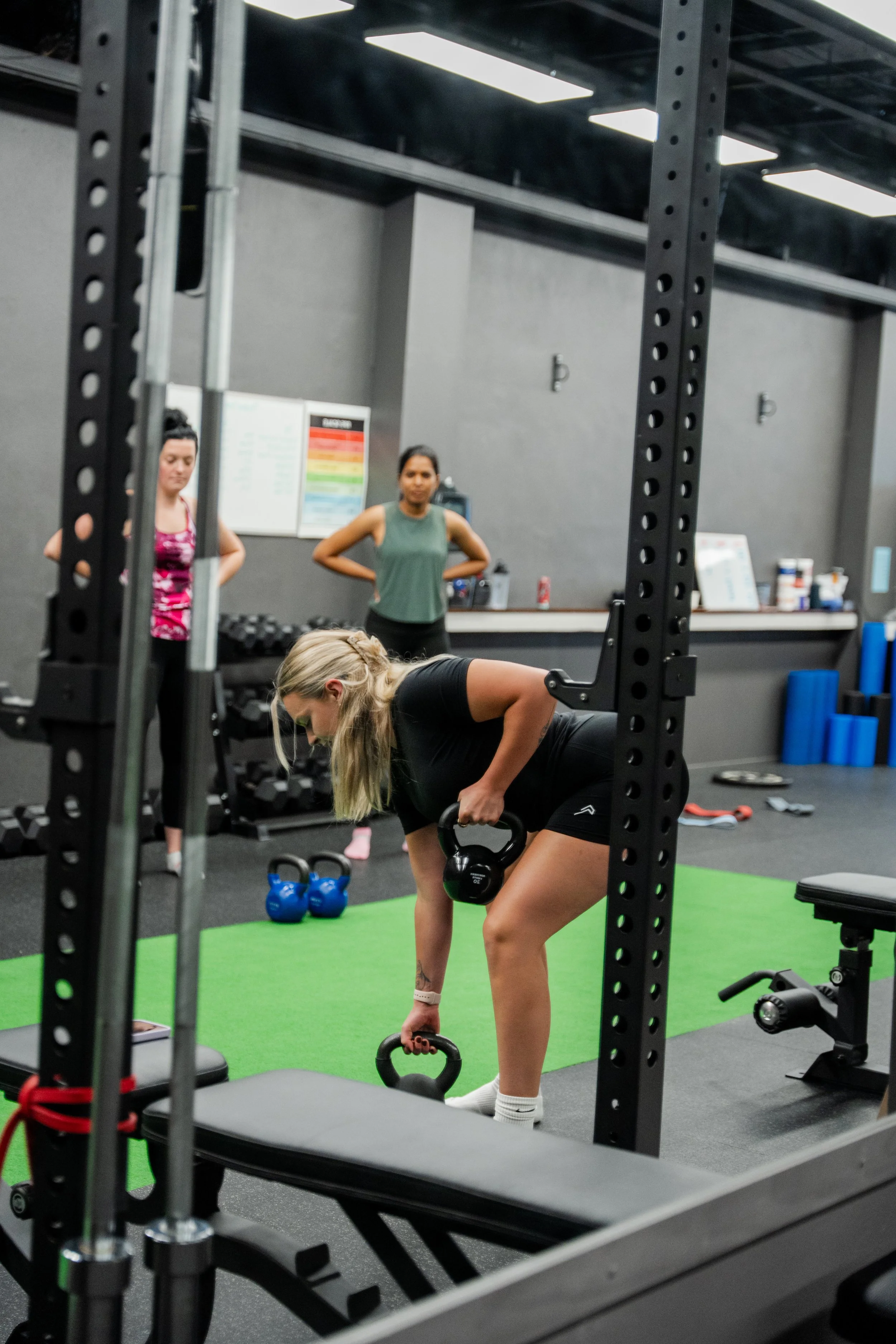A woman in black workout clothes lifts kettlebells in a gym with two women observing in the background.