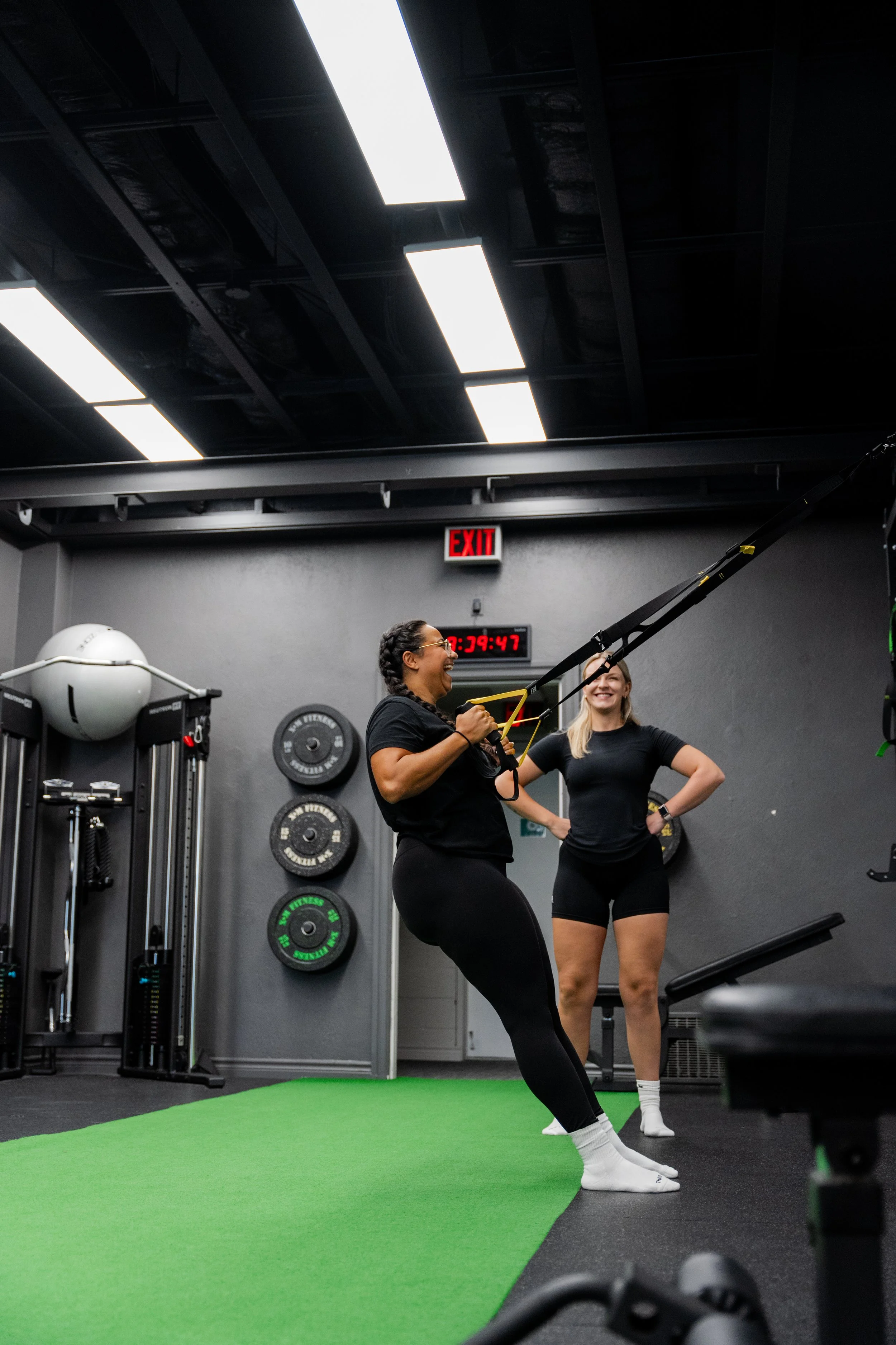 Two women exercising with resistance bands in a gym, one woman smiling while working out and the other woman standing behind her with hands on hips, on a green mat with gym equipment and weight plates in the background.