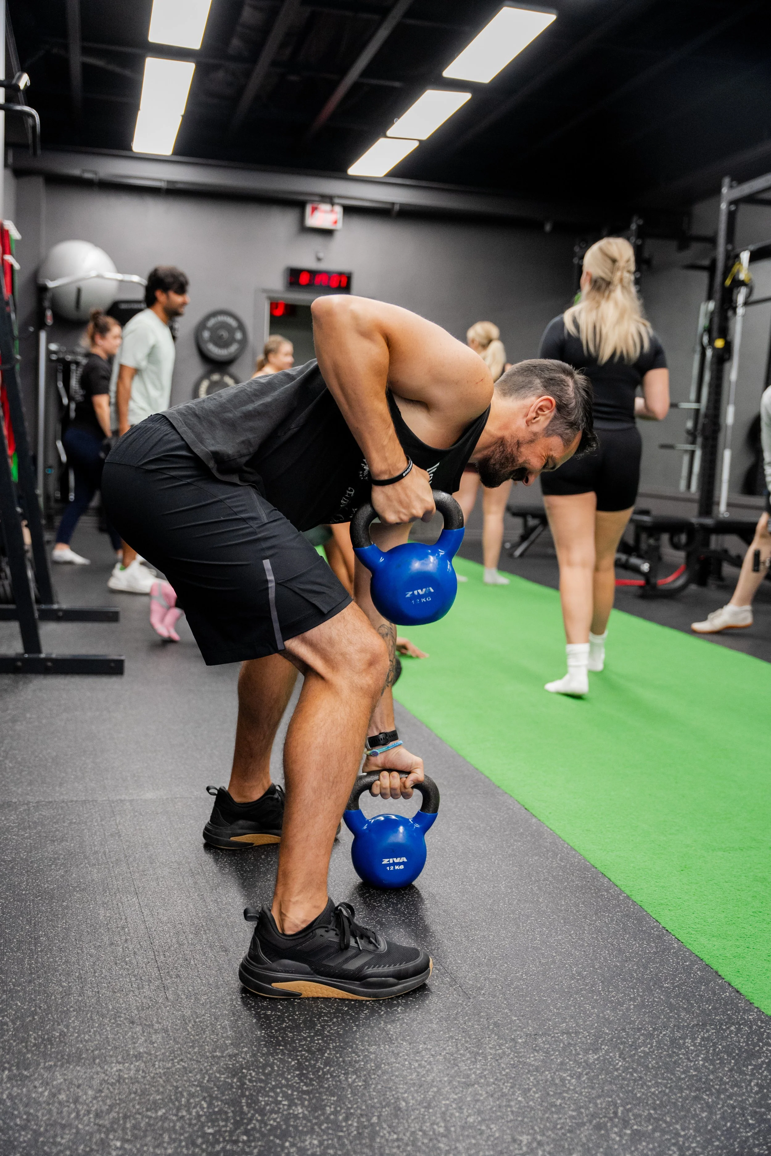 A man performing a workout with kettlebells in a gym, surrounded by other people exercising and gym equipment.
