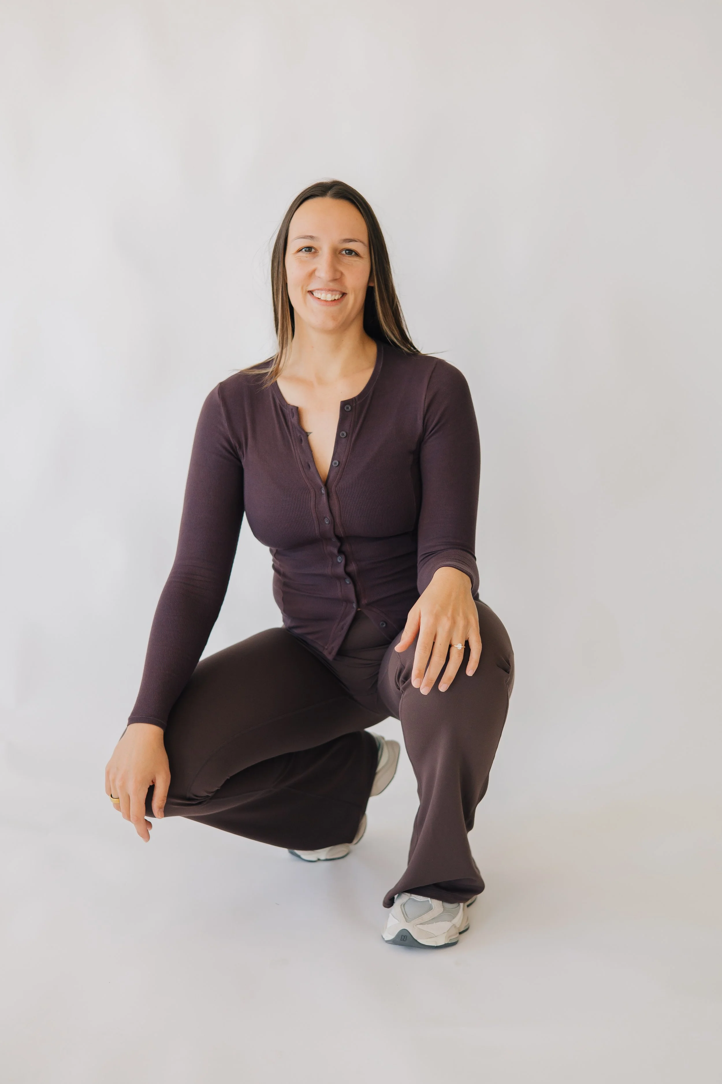 A woman kneeling on one knee with a cheerful expression, wearing a dark purple cardigan, dark pants, and white sneakers, against a plain light background.