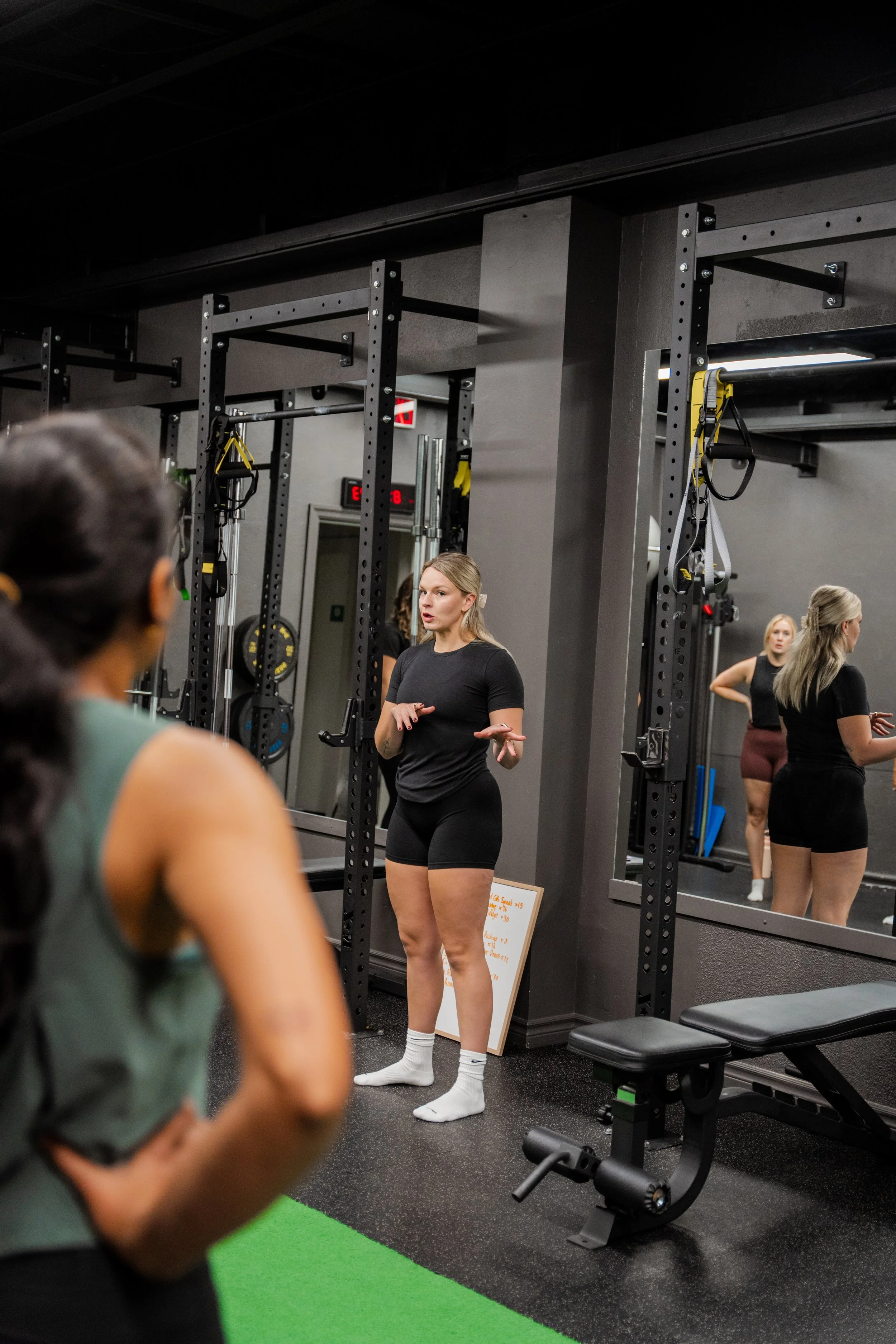 A fitness trainer giving instructions to a woman in a gym, with exercise equipment and mirrors on the wall in the background.