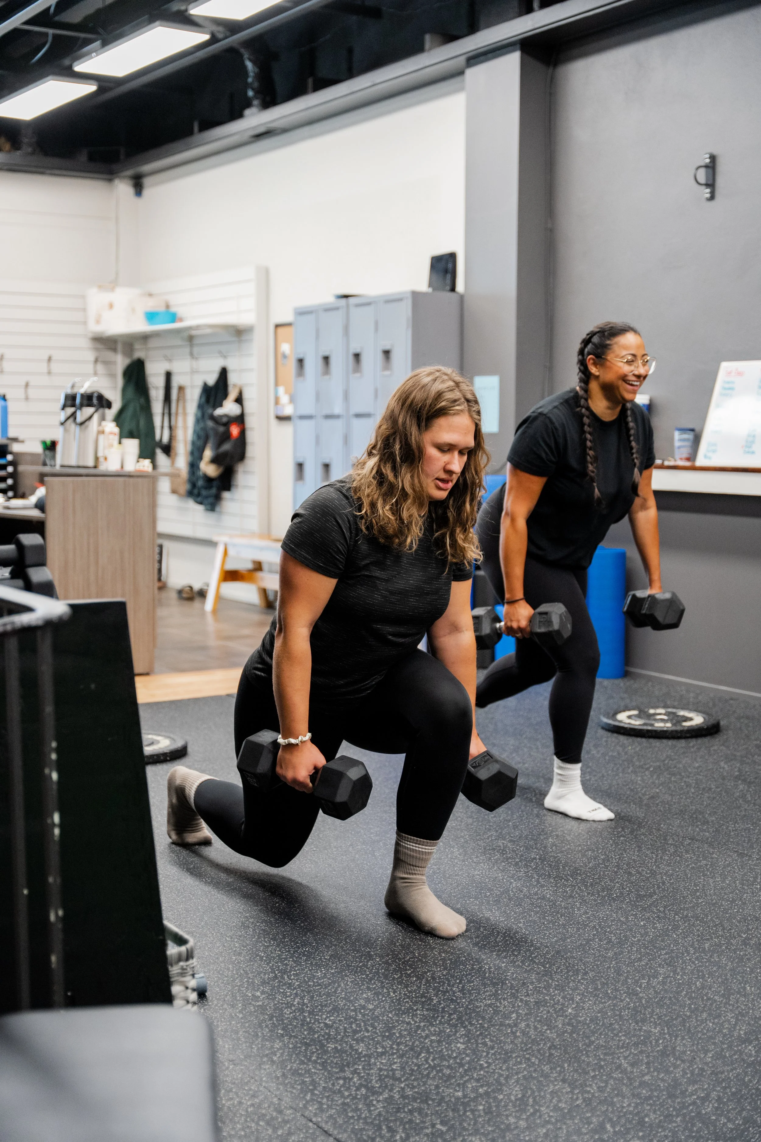 Two women lifting dumbbells while performing lunges in a gym. One woman is smiling, and the other appears focused.