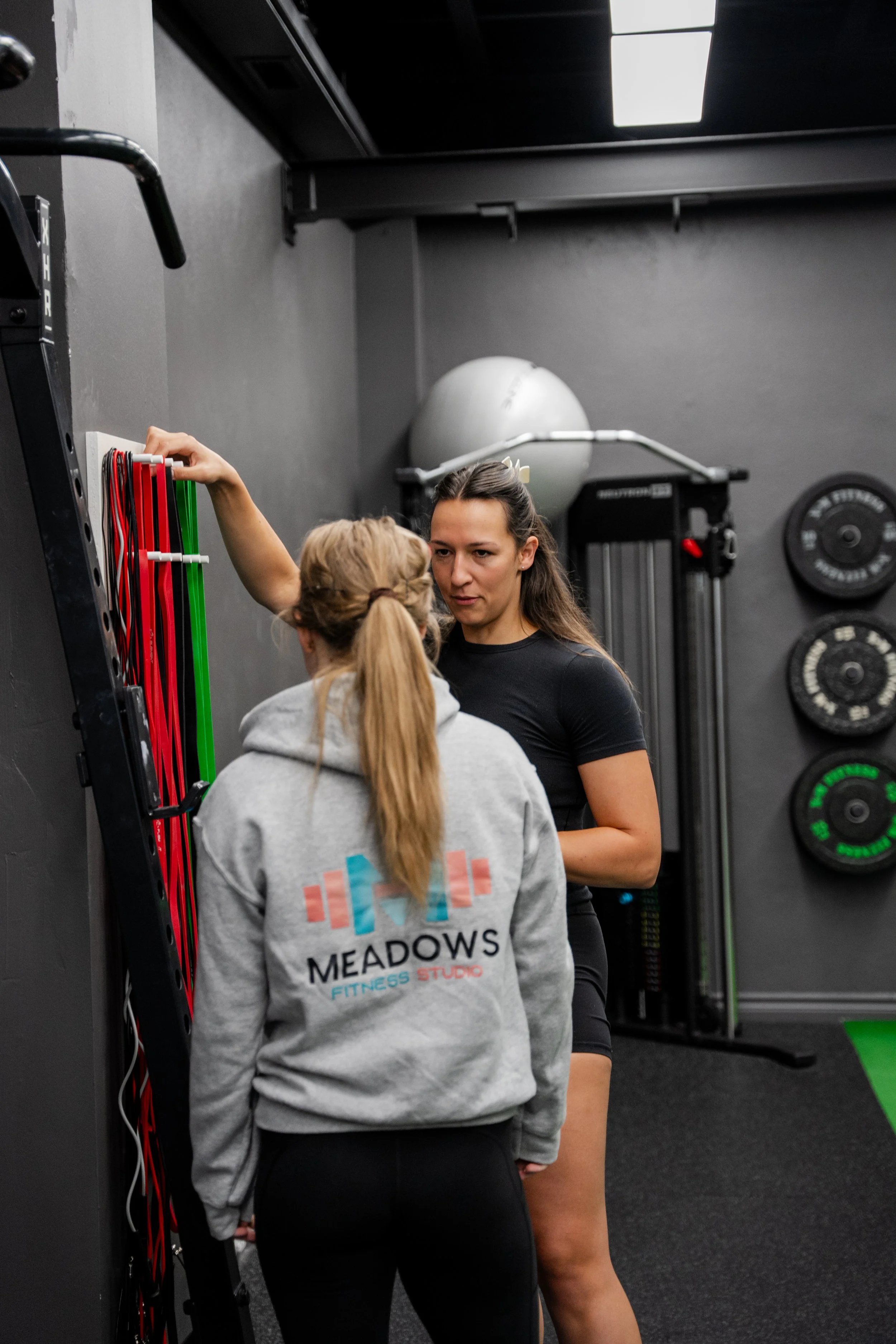 A female fitness instructor is giving instructions to a woman at a gym. The instructor, wearing a black shirt, is pointing to resistance bands. The woman, with a ponytail, is wearing a gray hoodie with "Meadows Fitness Studio" on the back.