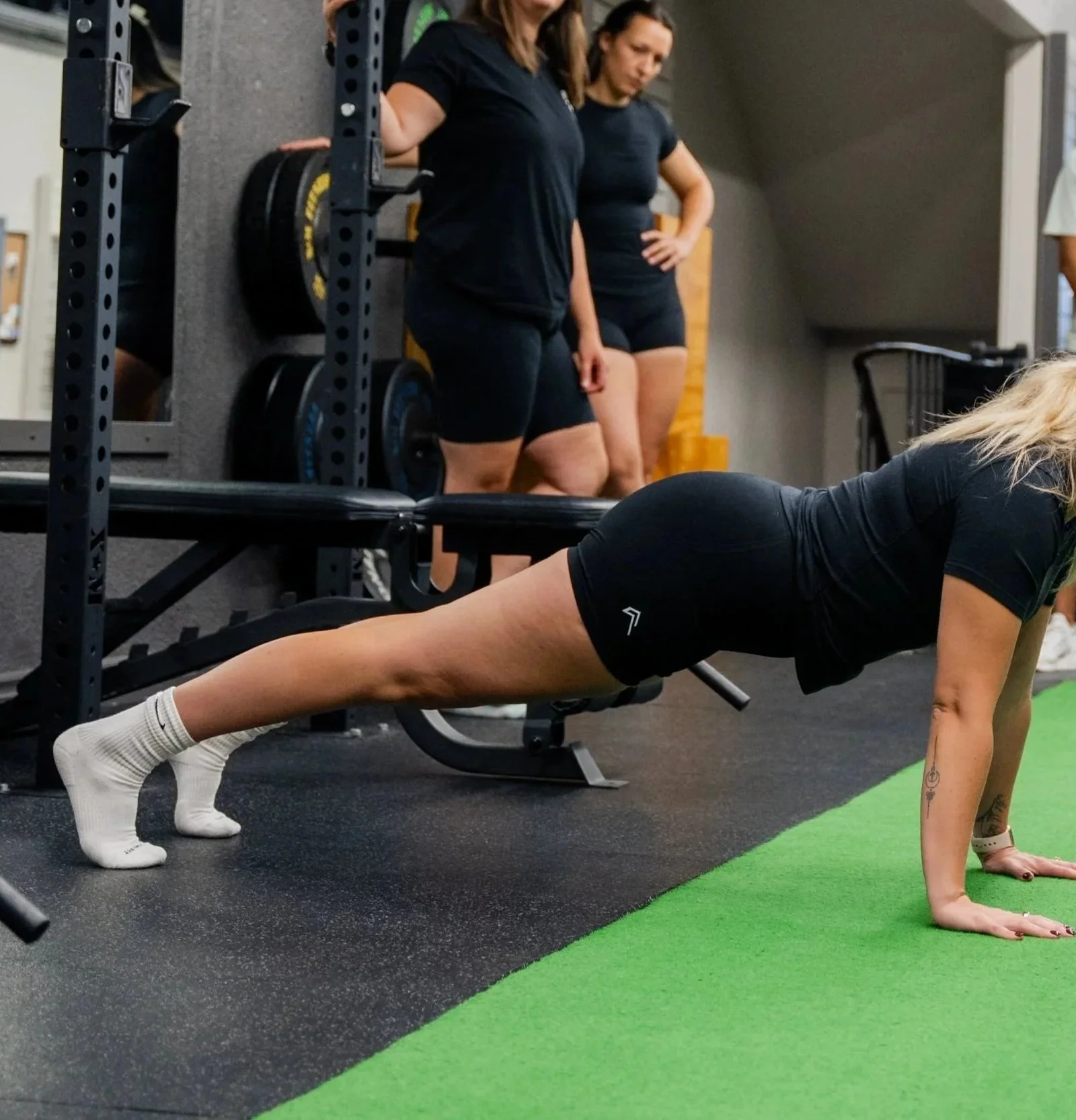 A woman in black workout clothes doing a plank exercise on a green mat in a gym, with two women observing in the background.