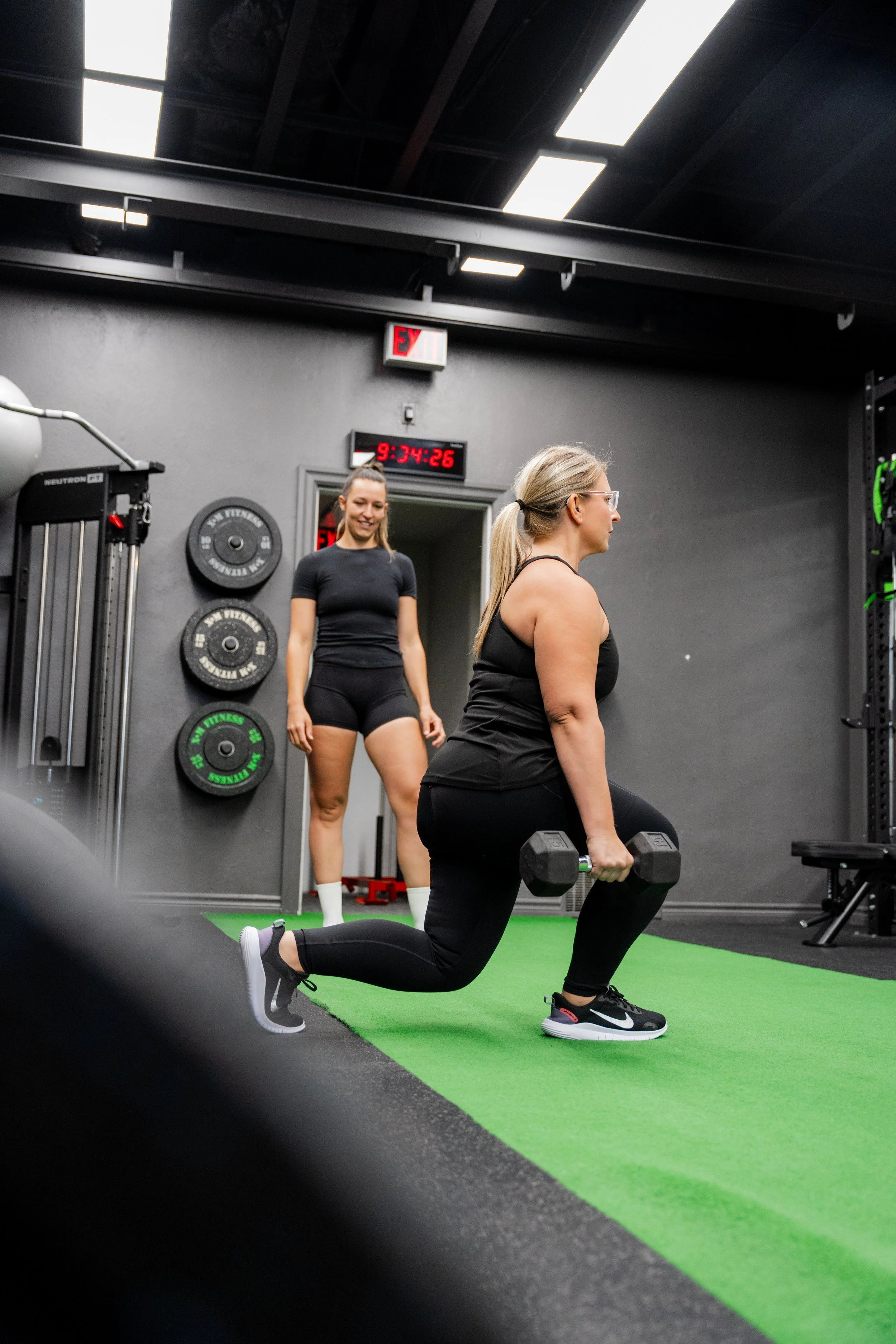 A woman exercising with dumbbells in a gym, with another woman standing nearby, and a digital clock on the wall showing the time 9:34:26.