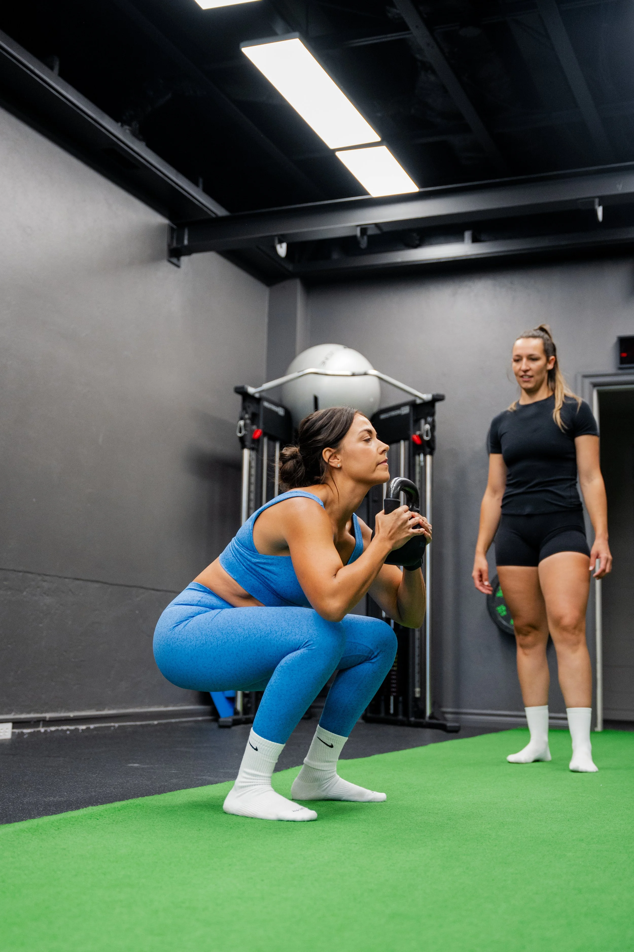 A woman in a blue workout outfit is performing a squat exercise with a kettlebell in a gym, supervised by another woman in black workout clothes.