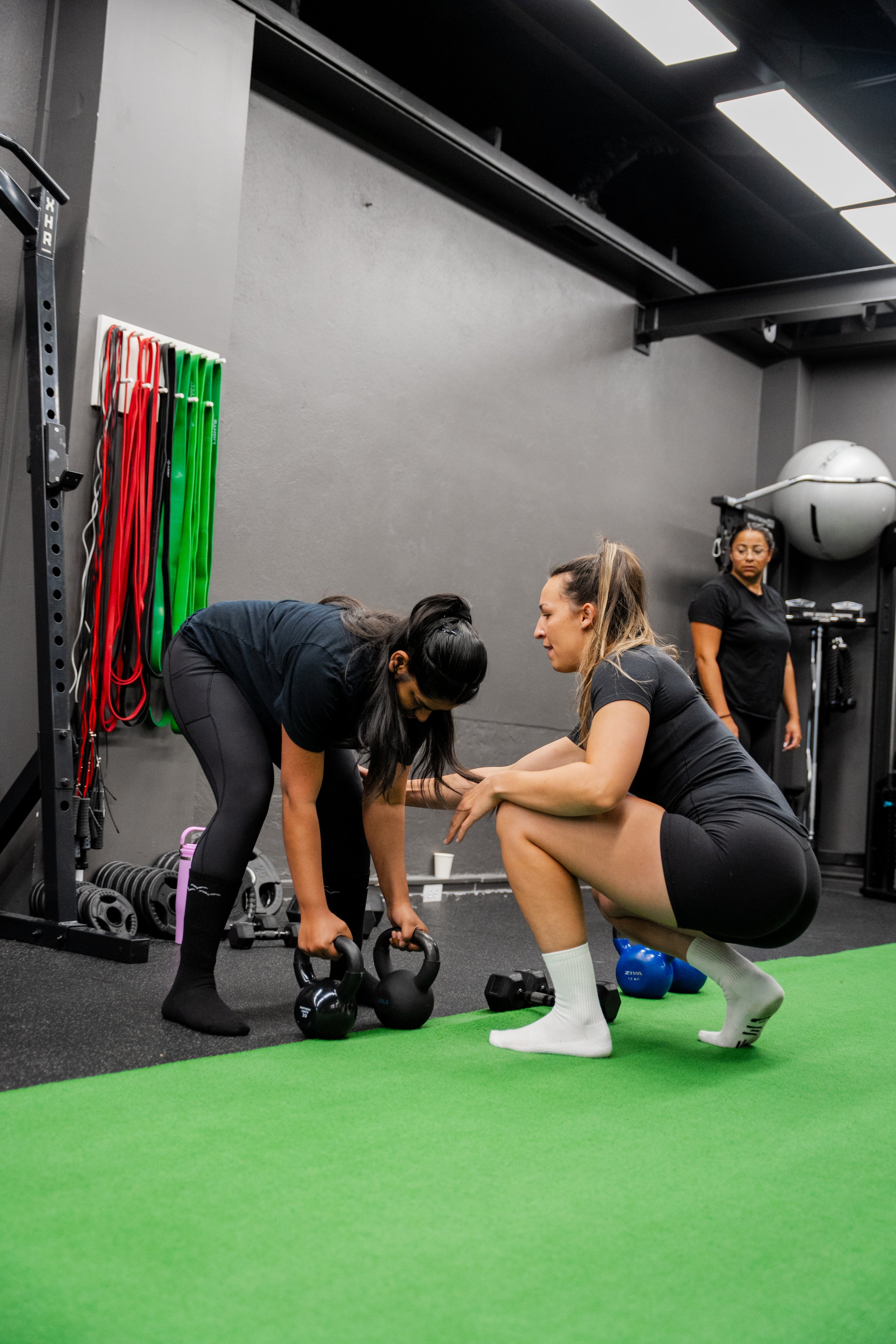 Two women exercising with kettlebells in a gym while a trainer assists, with workout equipment and resistance bands in the background.