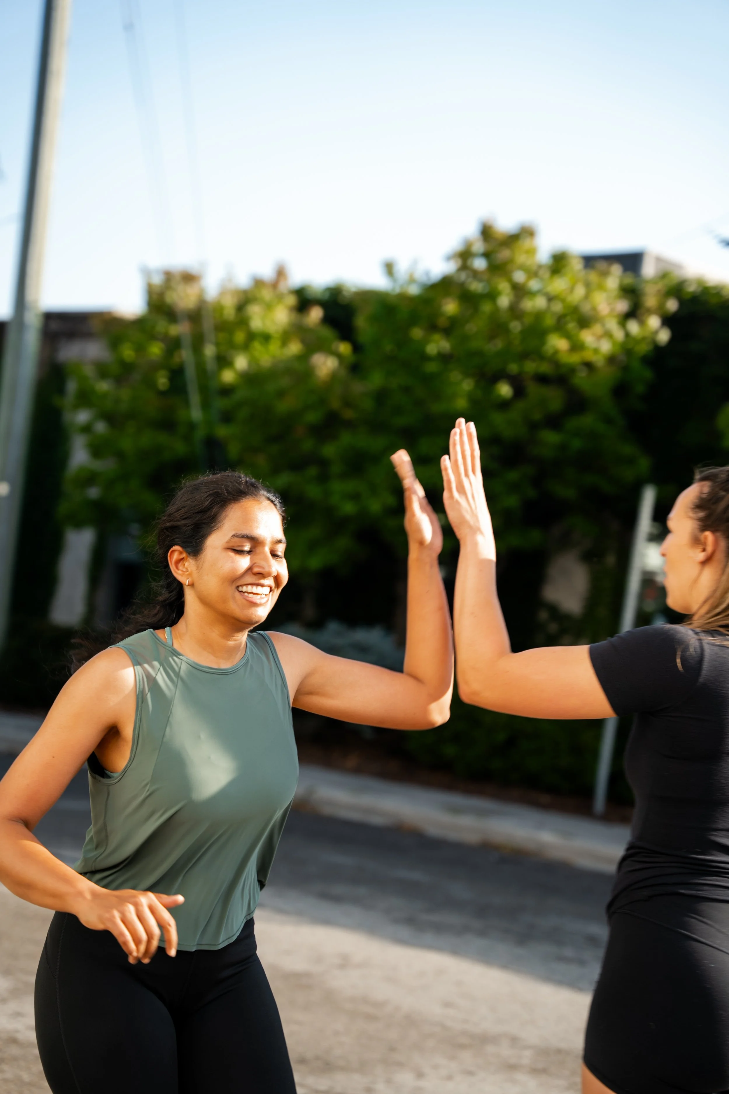 Two women in athletic clothing high-fiving outdoors in sunlight, with trees and a building in the background.
