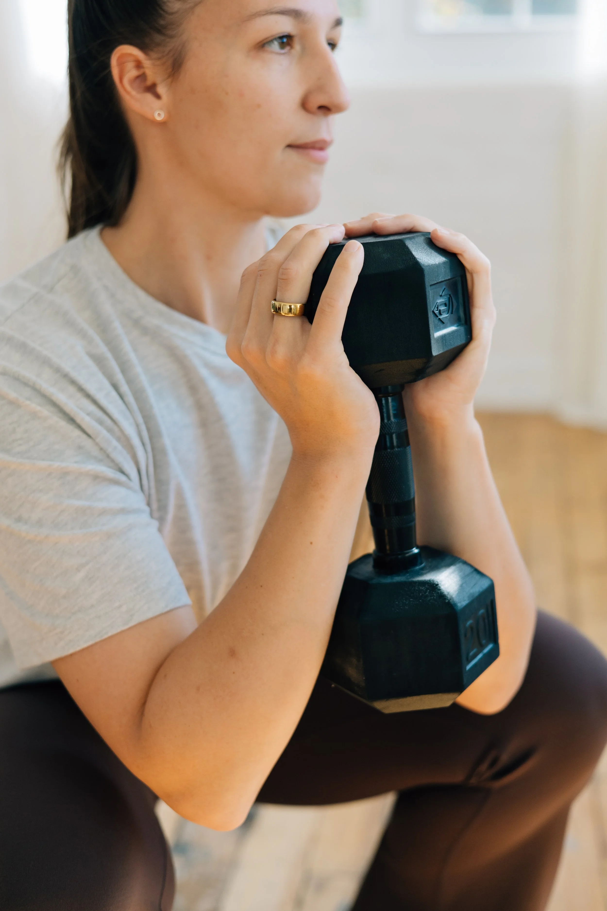 A woman sitting and holding a black dumbbell close to her chest in a gym or workout space.
