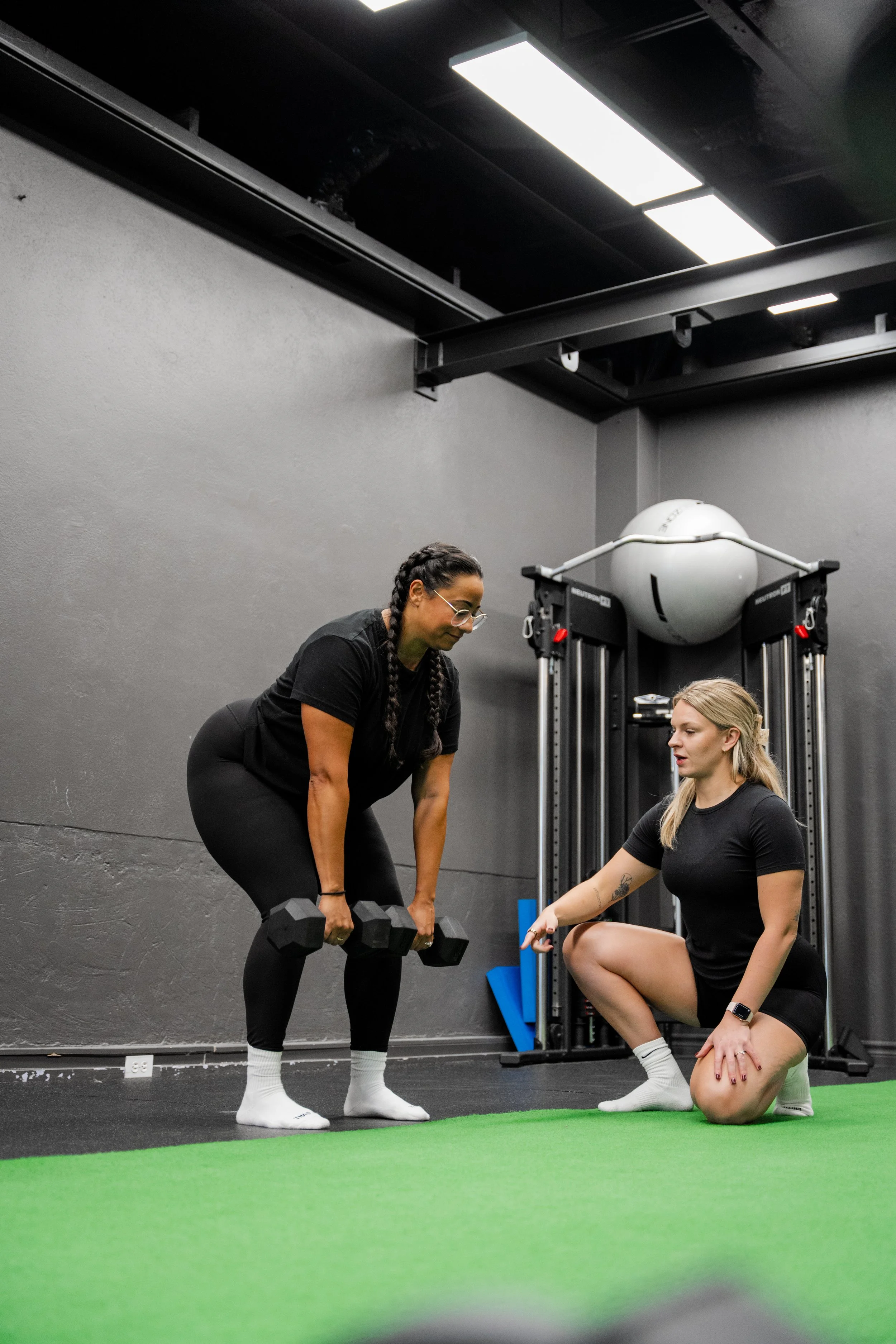 Two women in a gym, one standing and holding dumbbells, the other kneeling and giving instructions, with gym equipment in the background.