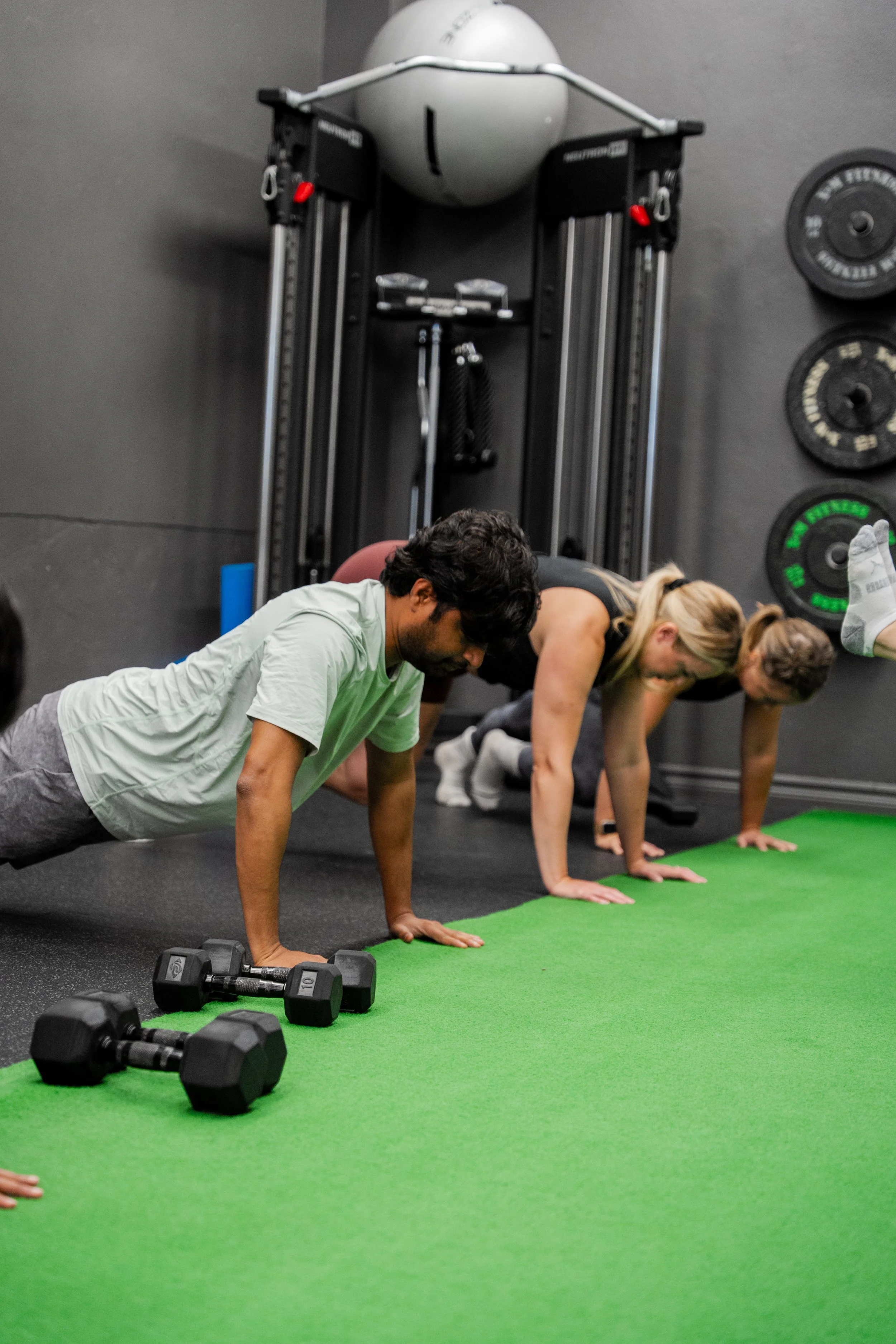 People doing push-ups on a green exercise mat in a gym, with dumbbells in the foreground and fitness equipment on the wall in the background.